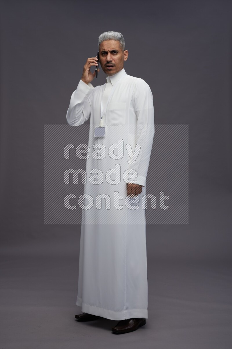 Saudi man wearing thob with neck strap employee badge standing talking on phone on gray background