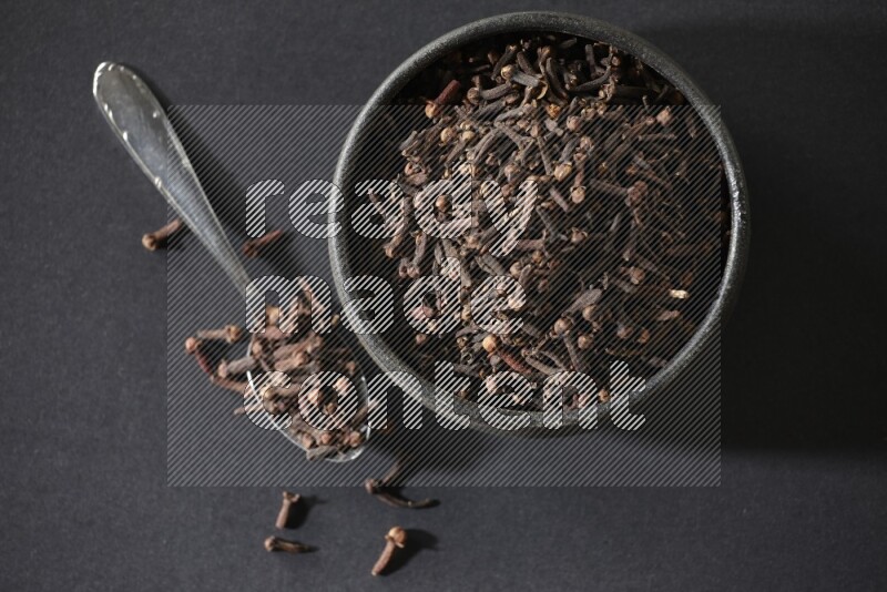 A black pottery bowl full of cloves and a metal spoon next to it on a black flooring
