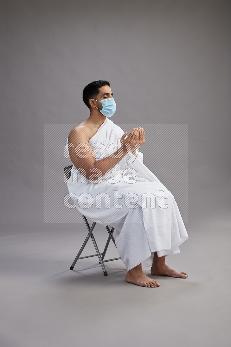 A man wearing Ehram with face mask sitting on chair dua'a on gray background