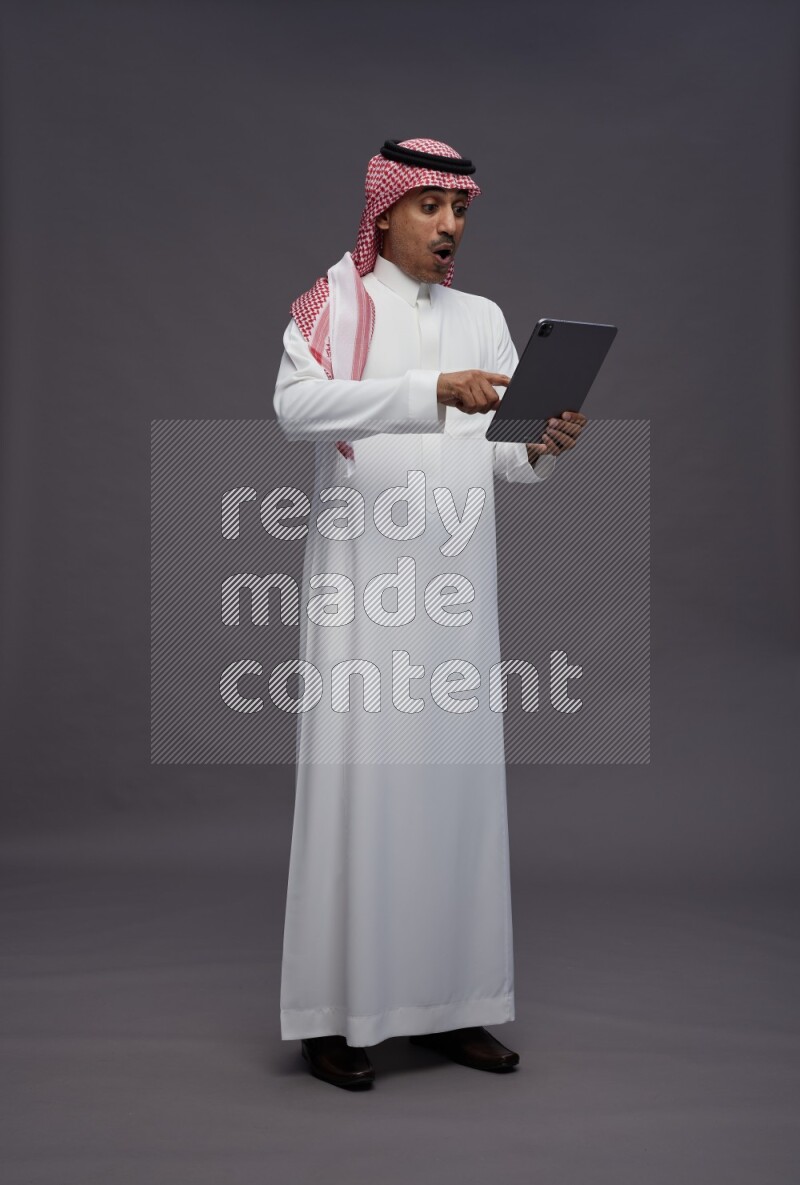 Saudi man wearing thob and shomag standing working on tablet on gray background