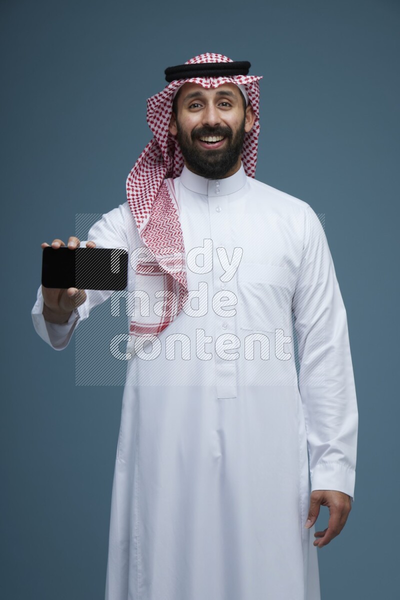 A man showing his phone Screen in a blue background wearing Saudi Thob with Shomag