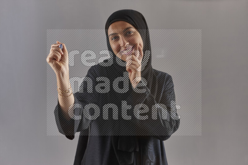 Woman in black abaya and hijab holding a marker pen to write on a transparent board with different reactions and expressions on grey background