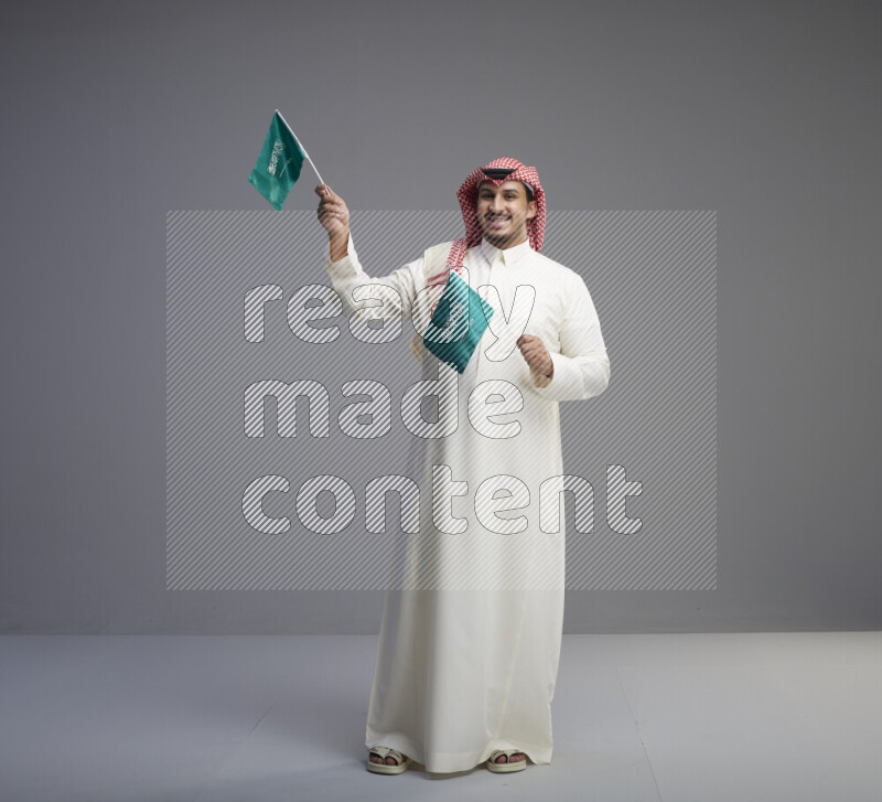 A Saudi man standing wearing thob and red shomag raising small Saudi flag on gray background