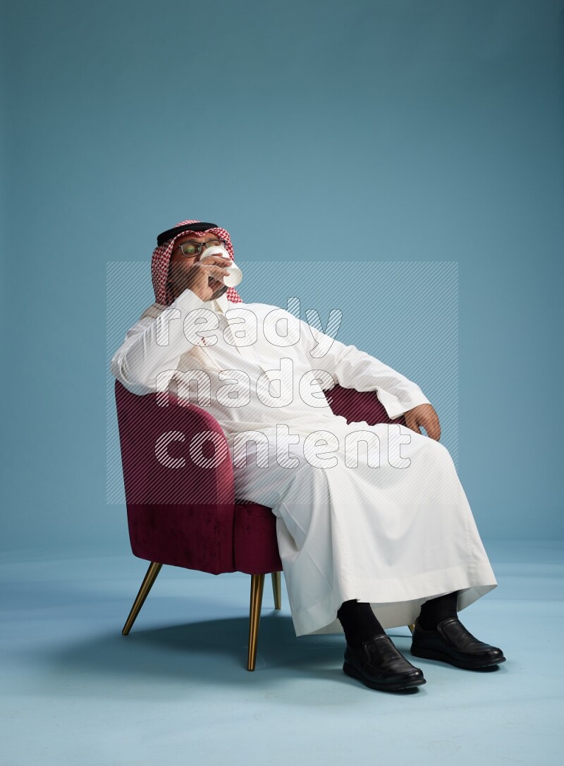 Saudi Man with shimag sitting on chair drinking coffee on blue background
