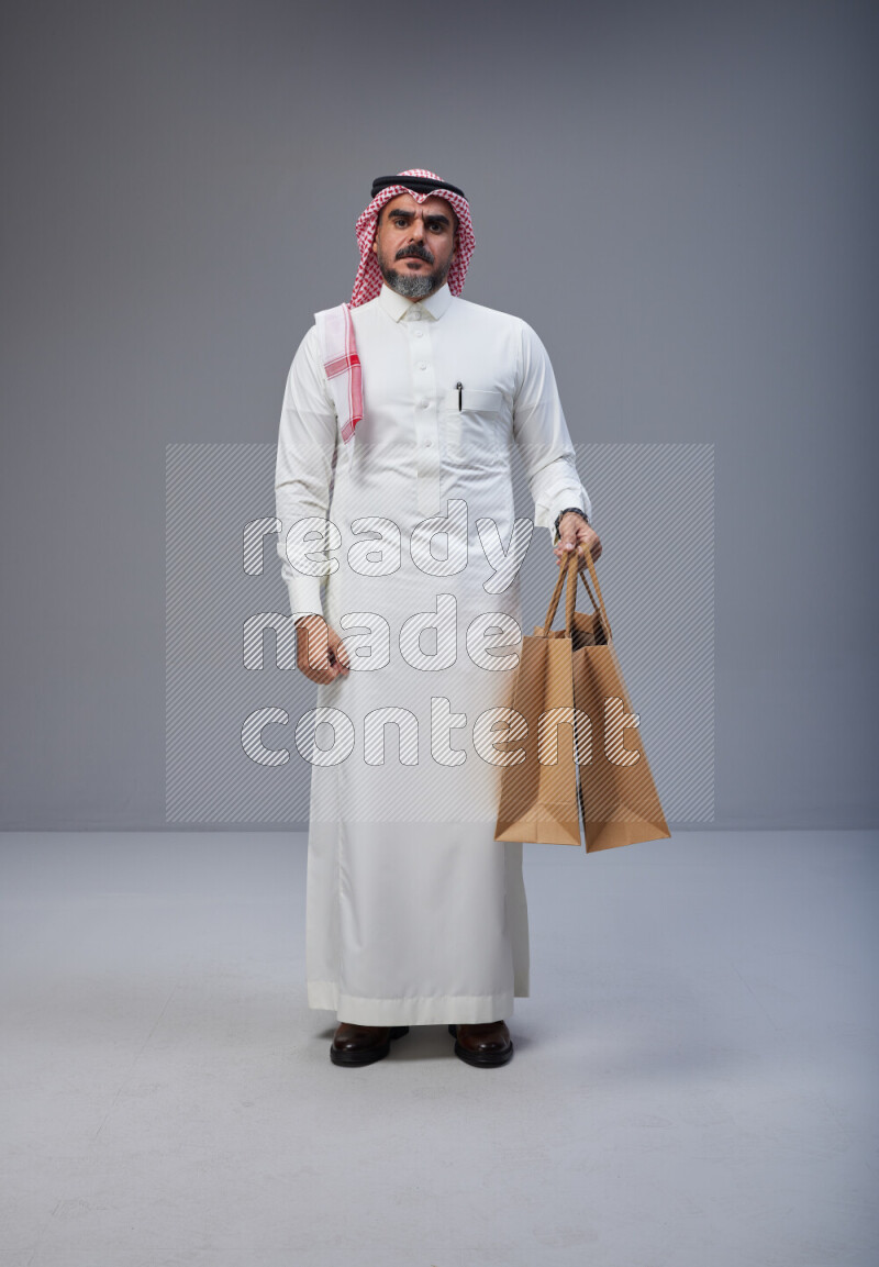 Saudi man Wearing Thob and red Shomag standing holding shopping bag on Gray background