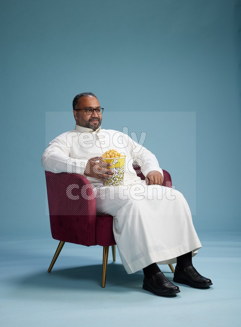 Saudi Man without shimag sitting on chair eating popcorn on blue background