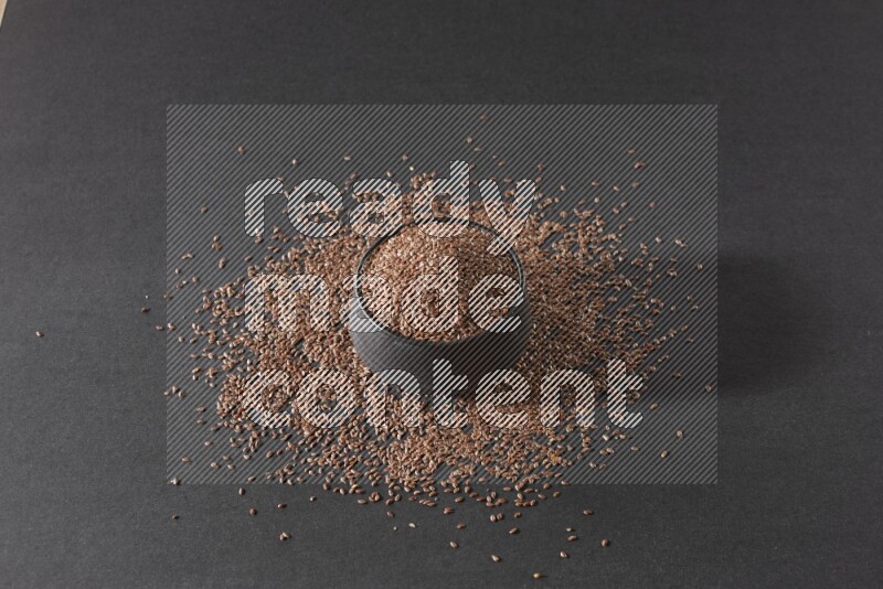 A black pottery bowl full of flaxseeds surrounded by the seeds on a black flooring