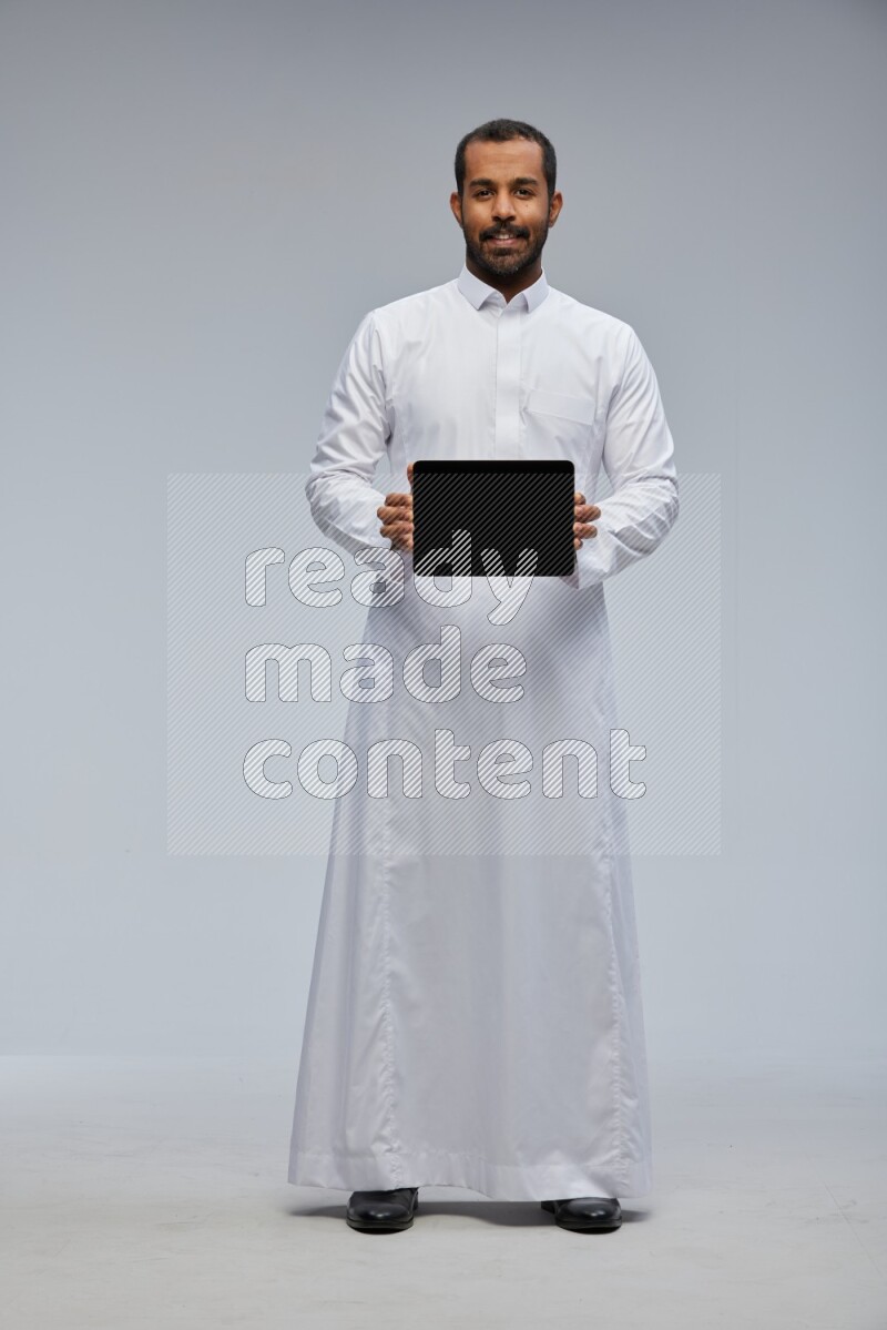 Saudi man wearing Thob standing showing tablet to camera on Gray background