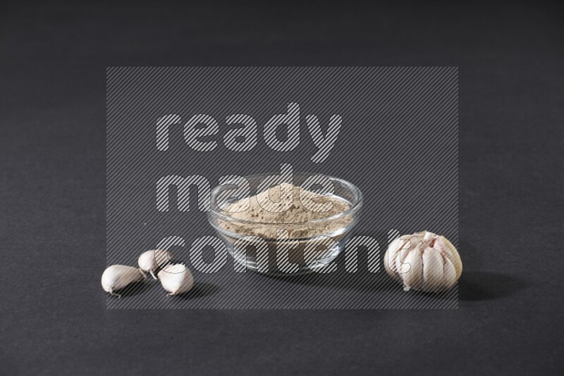 A glass bowl full of garlic powder with garlic bulb and some cloves beside it on a black flooring