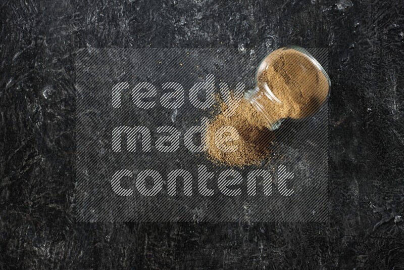 A flipped glass spice jar full of cumin powder and powder spilled out on a textured black flooring