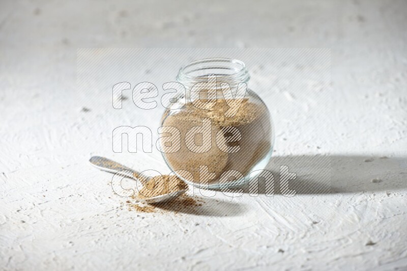 A glass spice jar and metal spoon full of cumin powder on textured white flooring