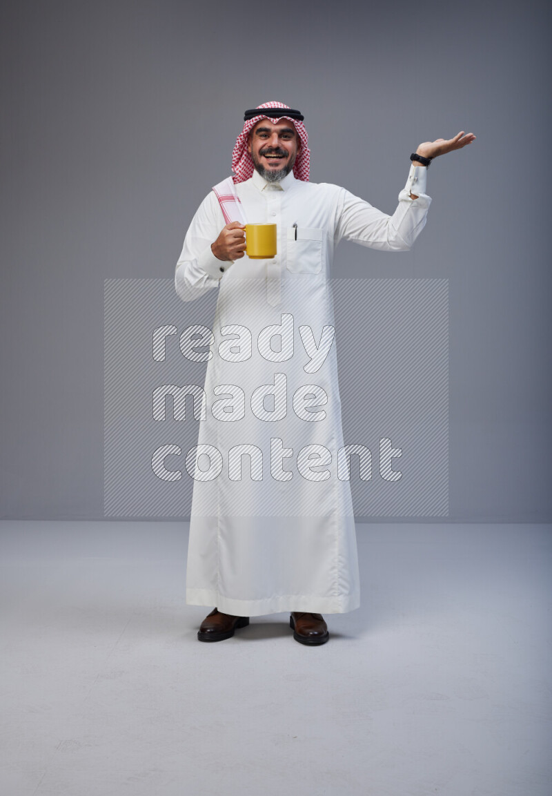 Saudi man Wearing Thob and red Shomag standing holding mug on Gray background