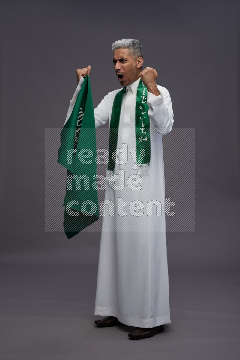 Saudi man wearing thob standing holding Saudi flag on gray background