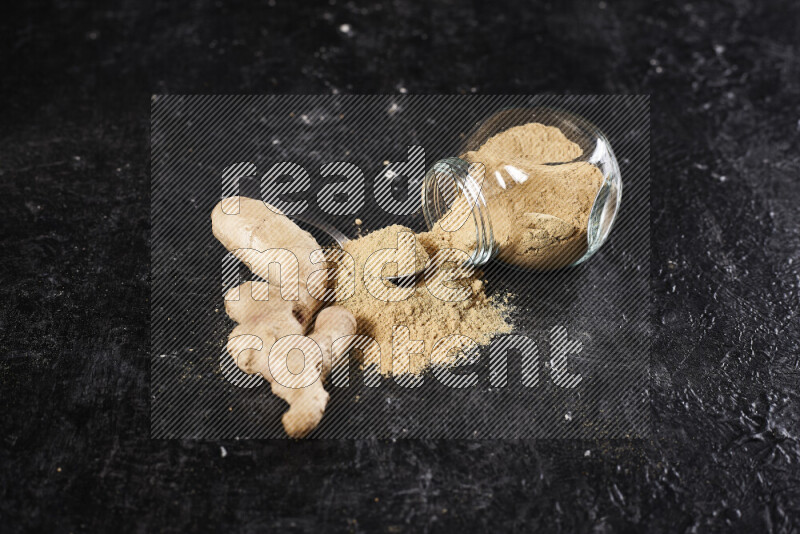 A glass jar full of ground ginger powder flipped with some spilling powder on black background