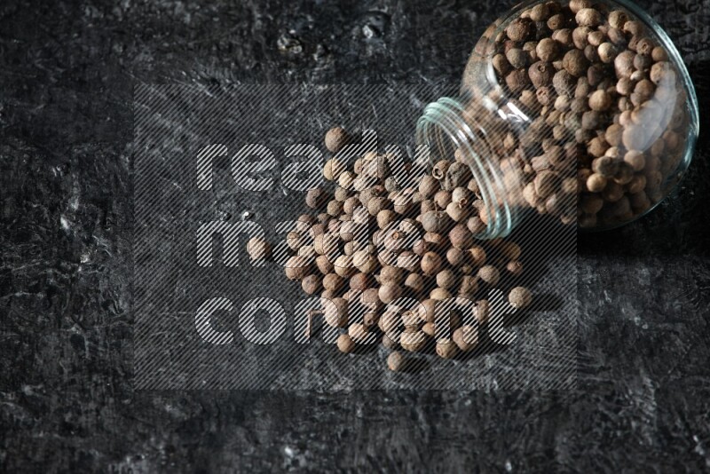 A flipped glass spice jar full of allspice whole balls and the balls spilled out of it on a textured black flooring