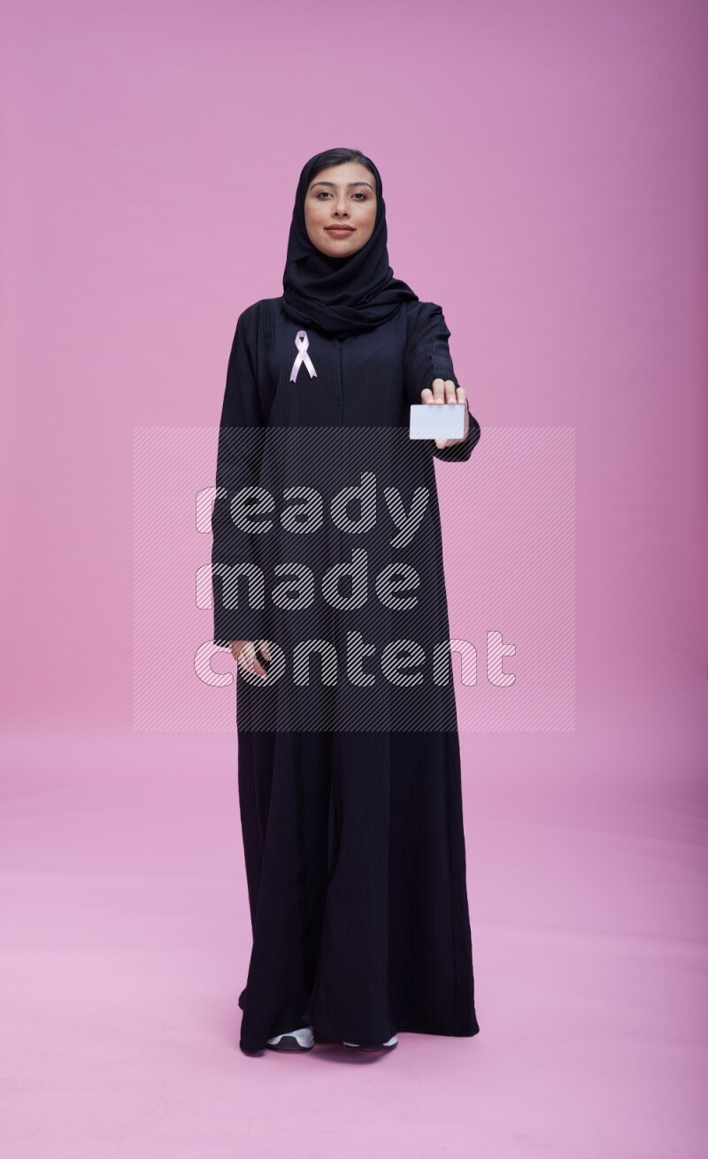 Saudi woman wearing pink ribbon on Abaya standing holding ATM card on pink background
