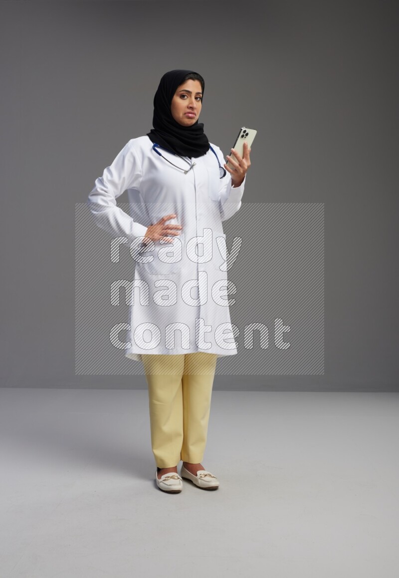 Saudi woman wearing lab coat with stethoscope standing texting on phone on Gray background