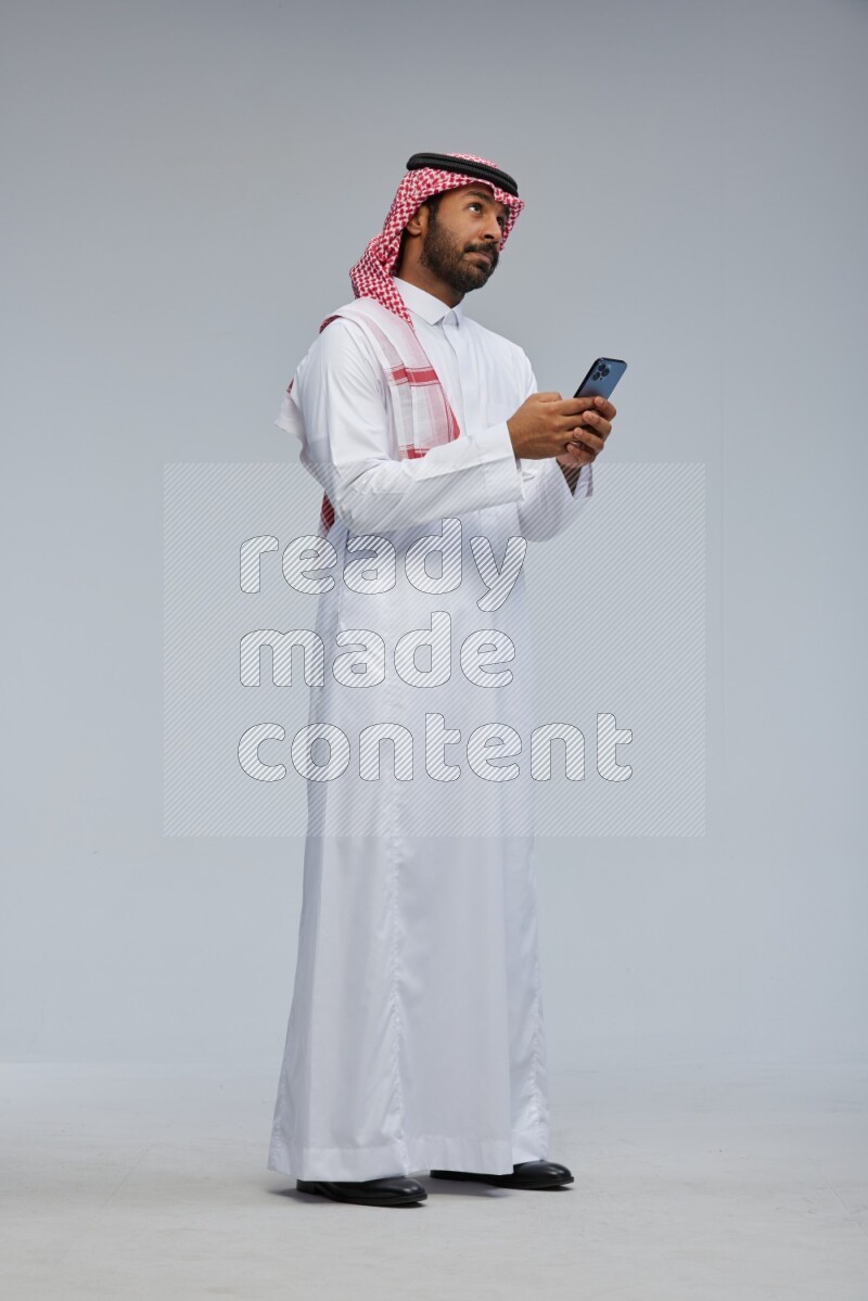 Saudi man Wearing Thob and shomag standing texting on phone on Gray background