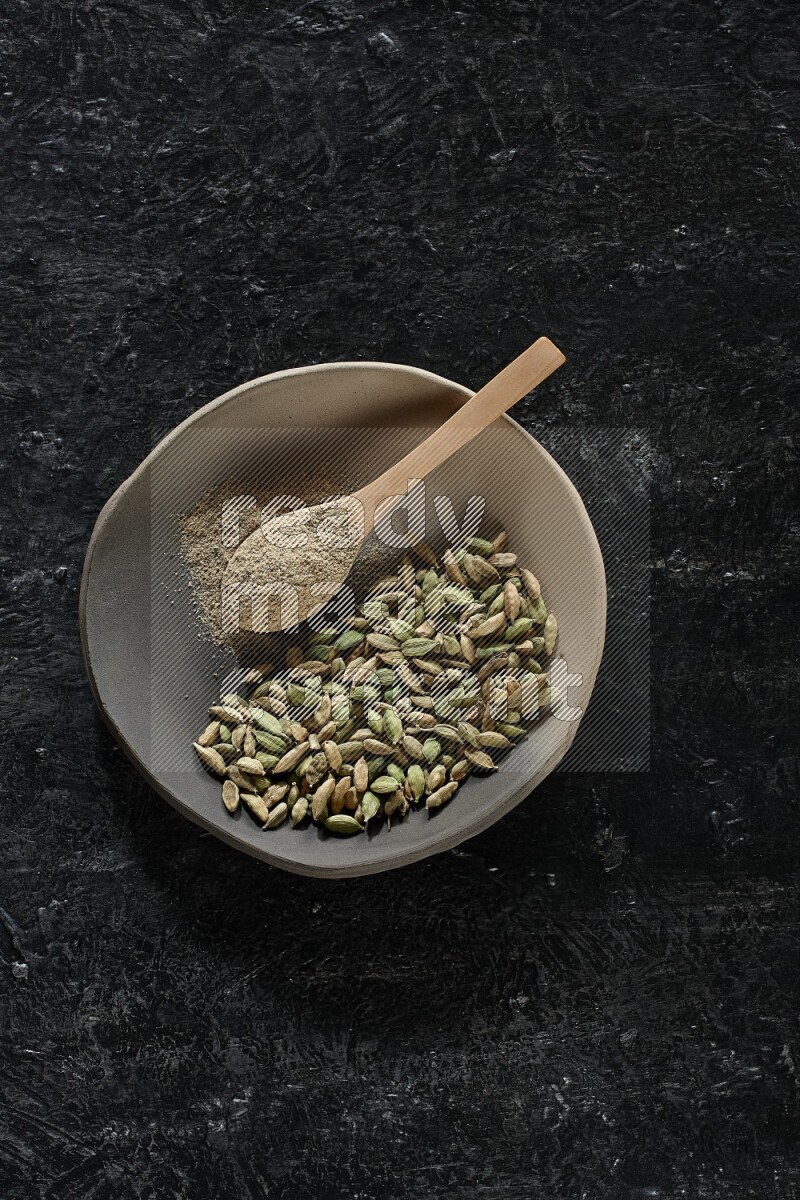 A plate filled with cardamom seeds and a wooden spoon full of cardamom powder on a textured black flooring