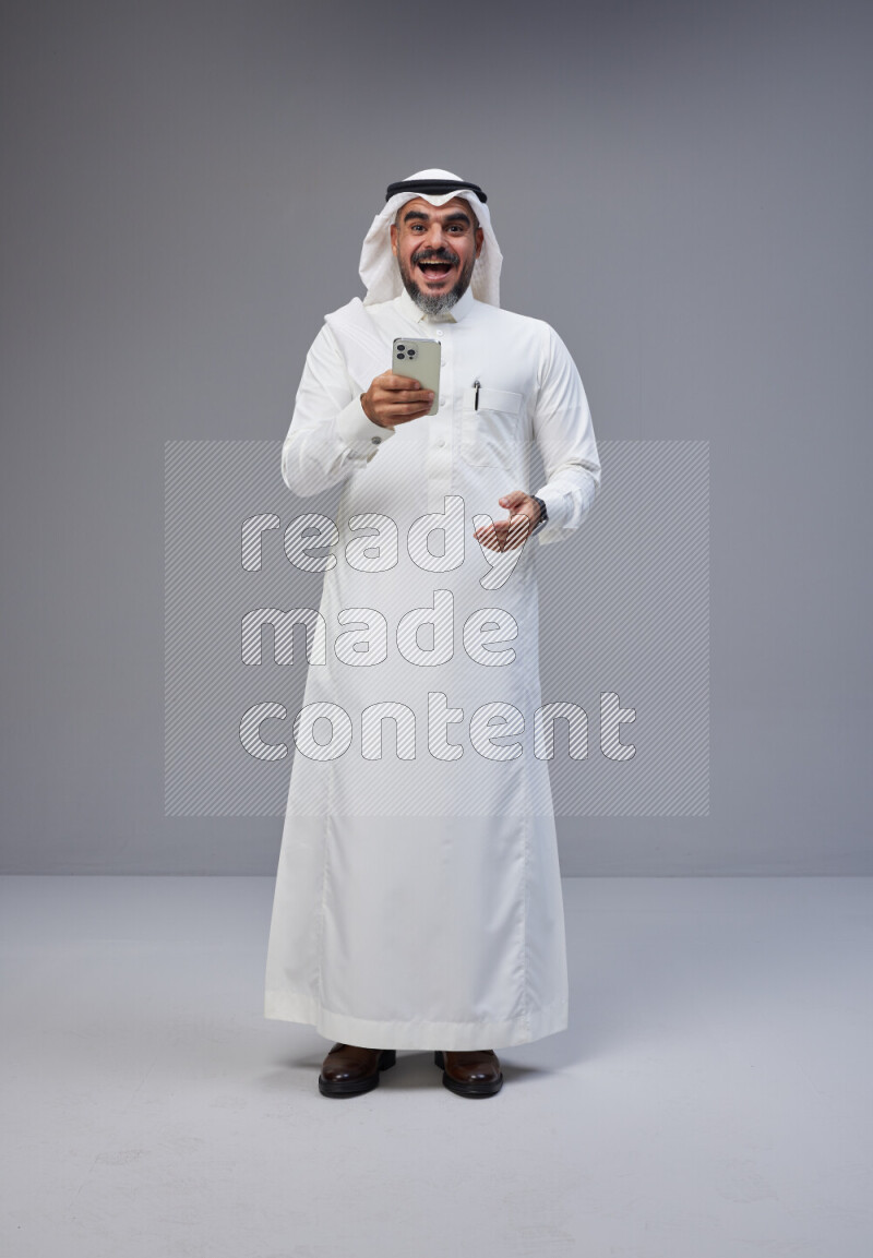 Saudi man Wearing Thob and white Shomag standing texting on phone on Gray background