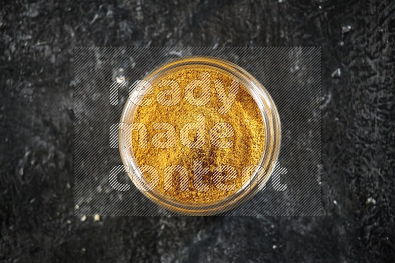 A glass jar full of turmeric powder on a textured black flooring