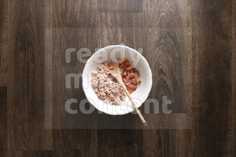 A white bowl full of tuna, colored bell pepper, sour cream, corn, parsley, black pepper powder and sauce, with wooden spoon on wooden background