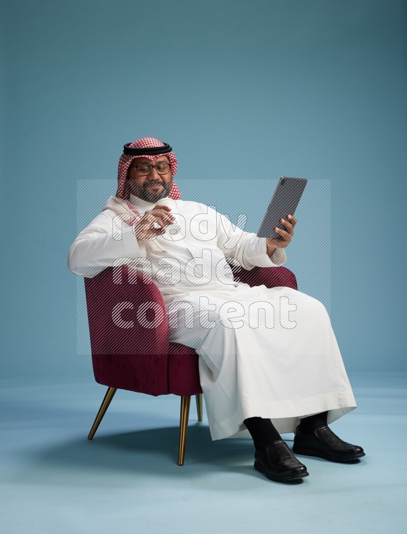 Saudi Man with shimag sitting on chair working on tablet on blue background