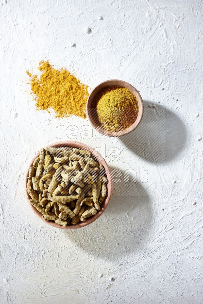 2 wooden bowls, one full of turmeric powder and the other full of dried turmeric whole fingers on a textured white flooring