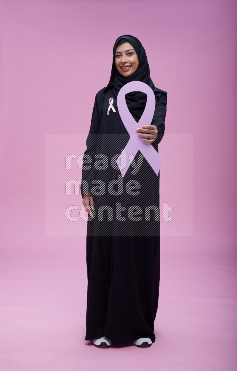 Saudi woman wearing pink ribbon on abaya standing holding awareness ribbon on plain pink background