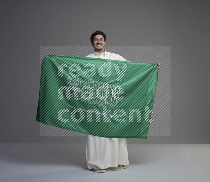 A Saudi man standing wearing thob holding big Saudi flag on gray background