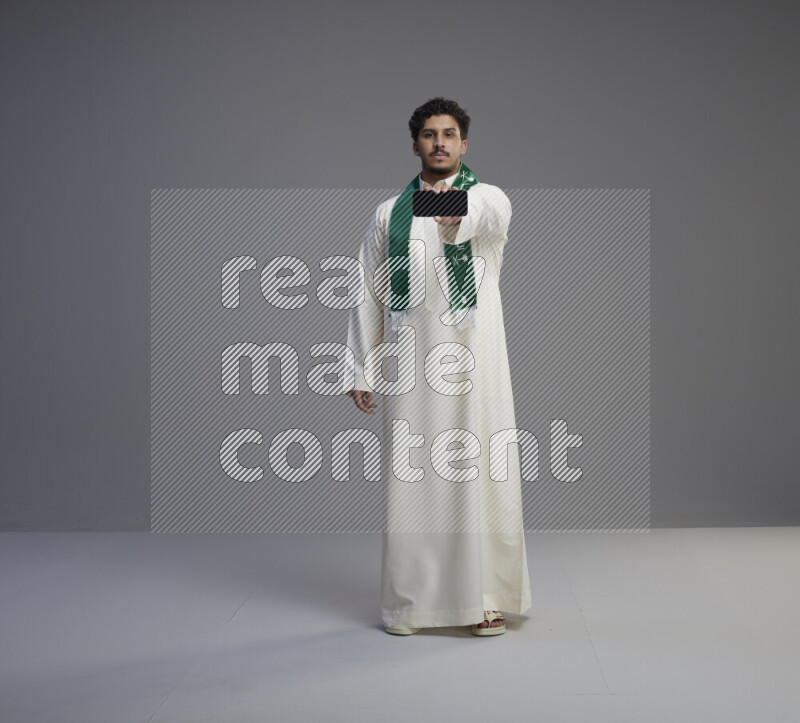 A Saudi man standing wearing thob and Saudi flag scarf showing phone to camera on gray background