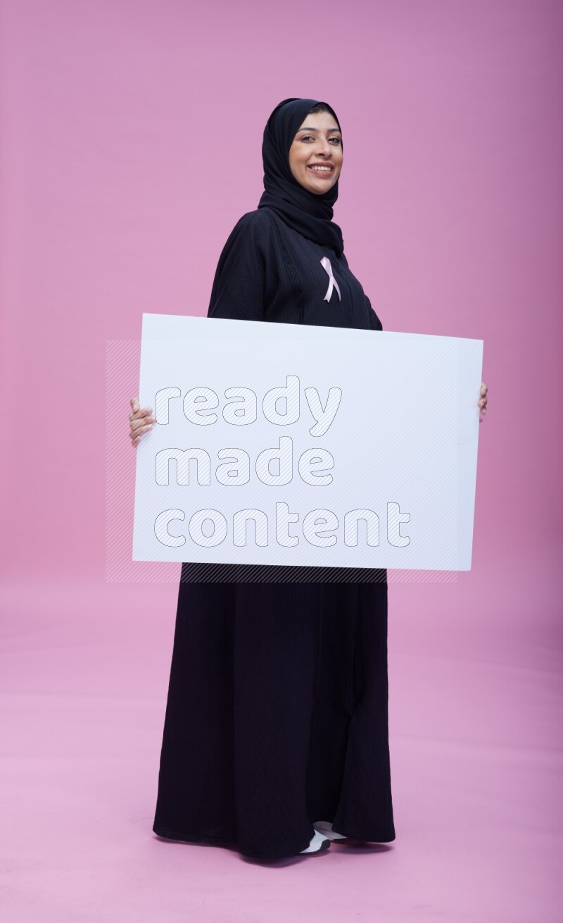 Saudi woman wearing pink ribbon on Abaya standing holding board on pink background