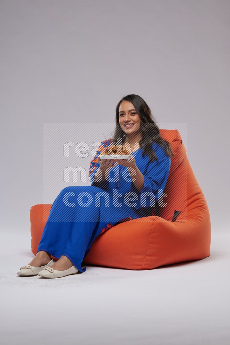 A Woman sitting on an orange beanbag wearing Jalabeya holding a plate of dates
