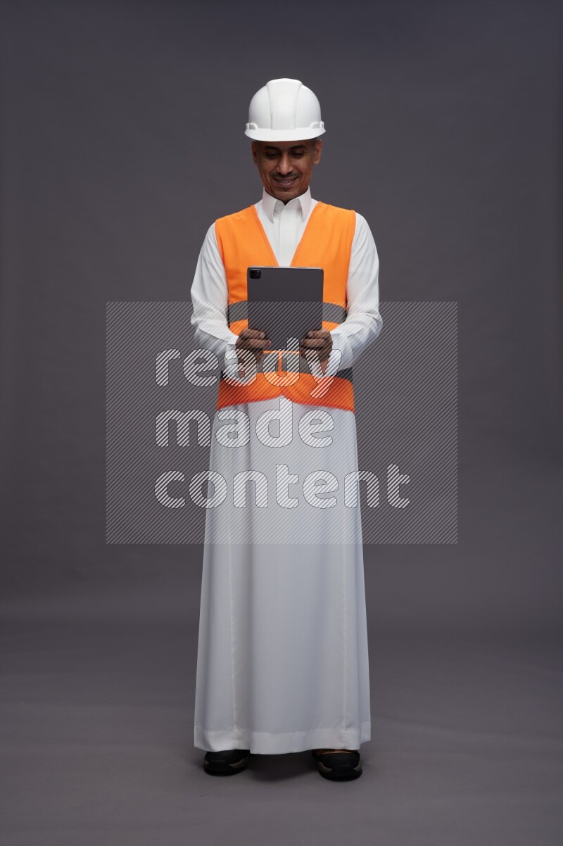 Saudi man wearing thob with engineer vest standing working on tablet on gray background