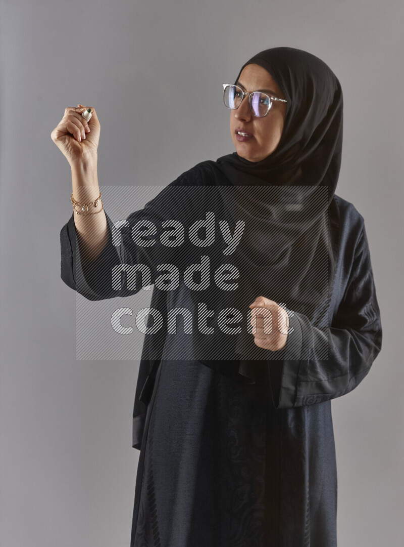 Woman in black abaya, hijab and glasses holding a marker pen to write on a transparent board with different reactions and expressions on grey background