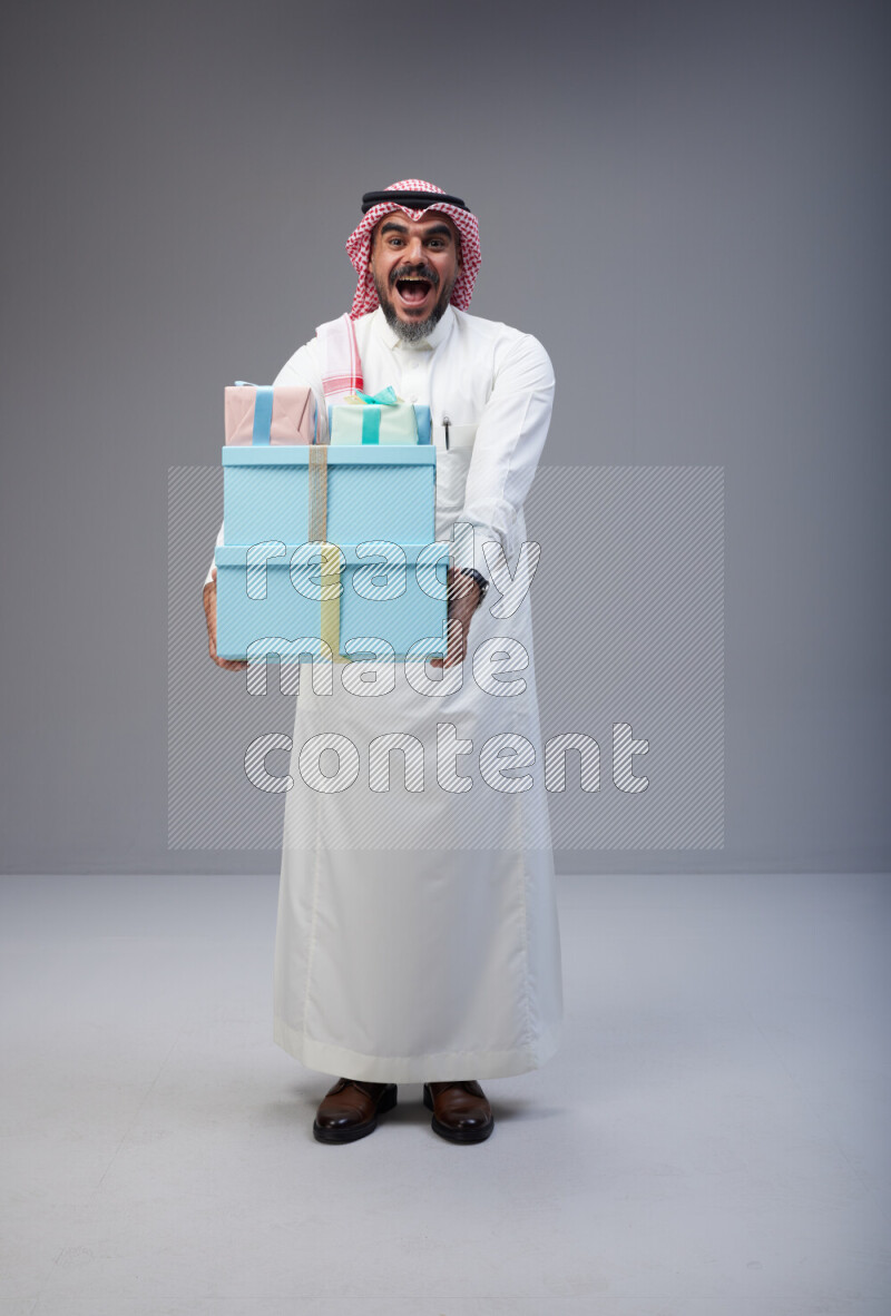 Saudi man Wearing Thob and red Shomag standing holding gift box on Gray background