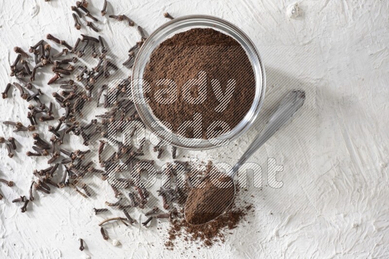 A glass bowl and a metal spoon full of cloves powder with cloves grains spread on a textured white flooring