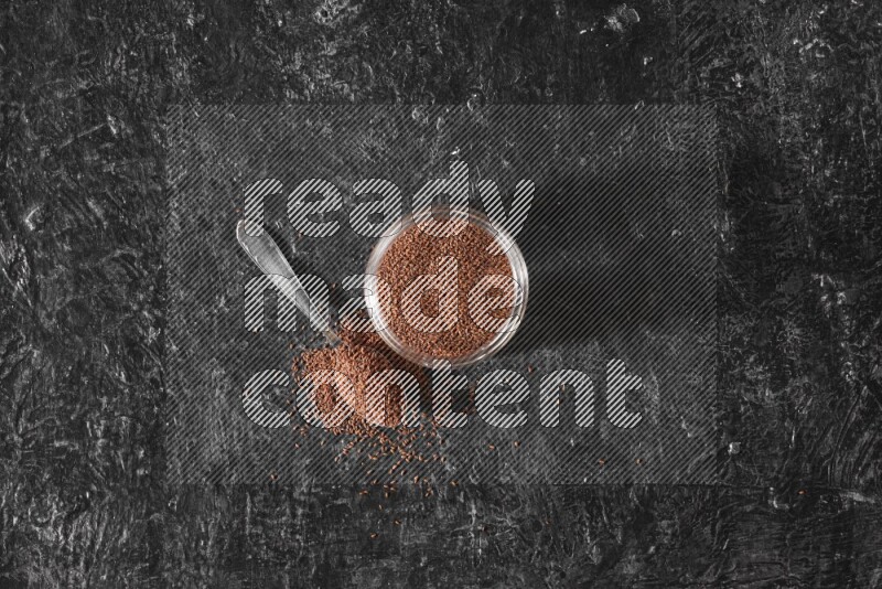 A glass jar full of garden cress seeds with a metal spoon full of the seeds on a textured black flooring