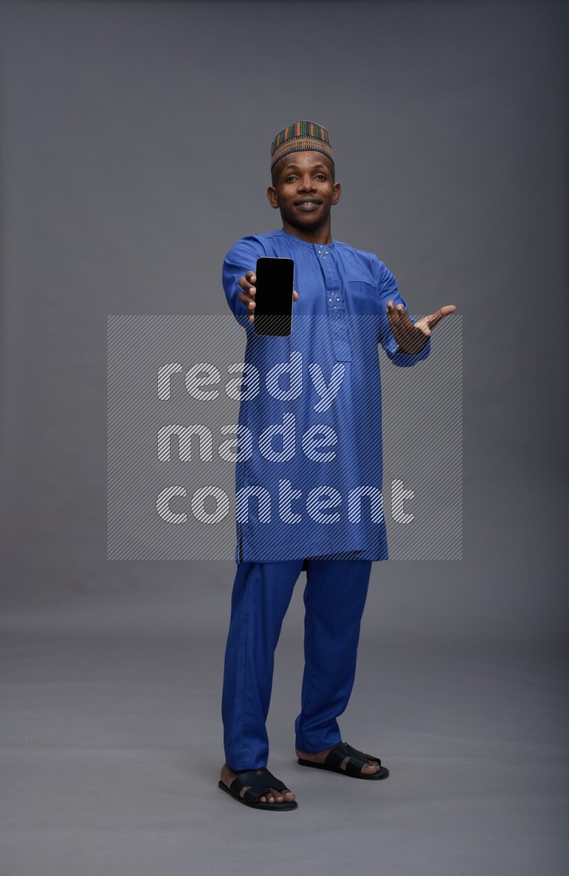 Man wearing Nigerian outfit standing showing phone to camera on gray background