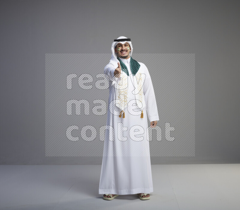 A Saudi man standing wearing thob and white shomag with face painting and Saudi flag scarf on gray background