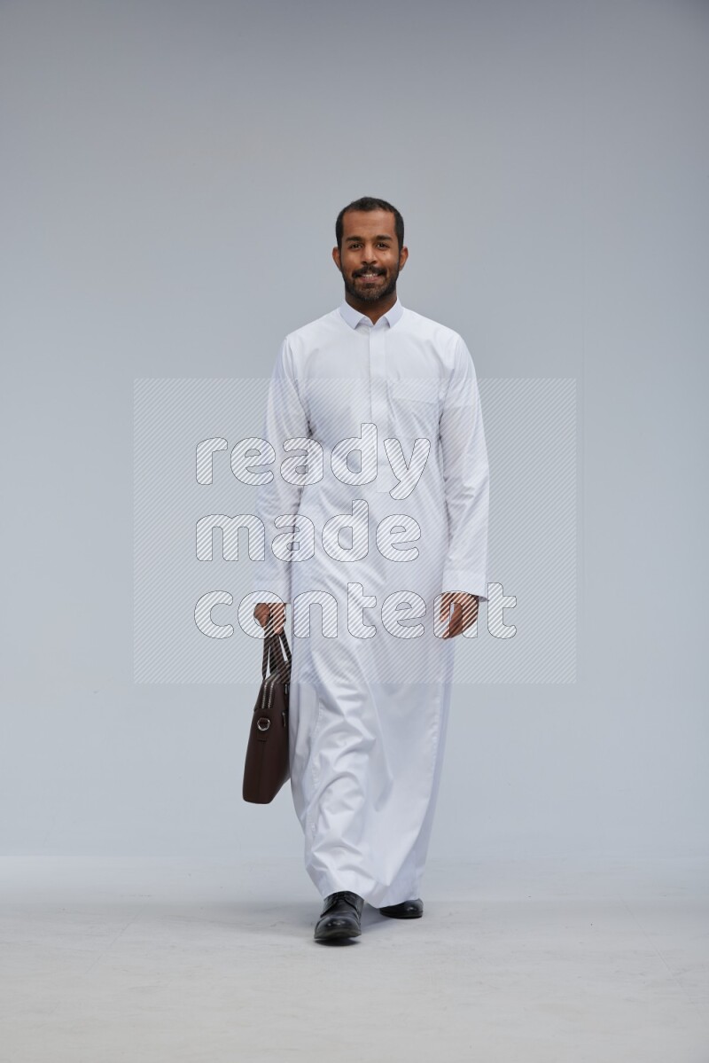 Saudi man wearing thob standing holding suitcase on gray background