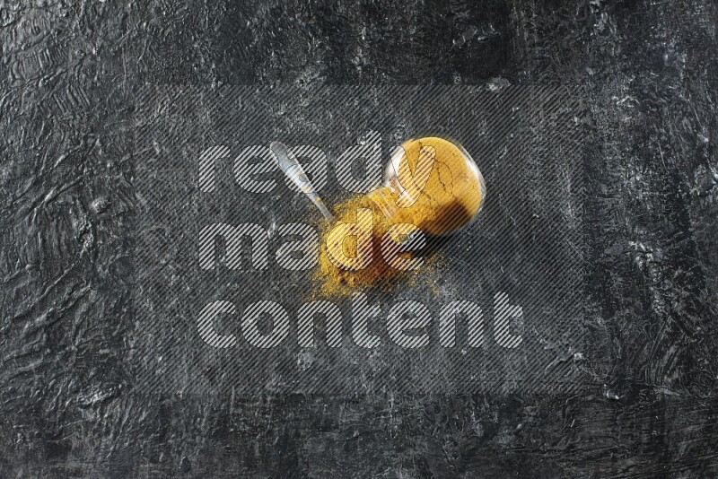 A flipped glass spice jar and a metal spoon full of turmeric powder and powder spilled out of it on textured black flooring