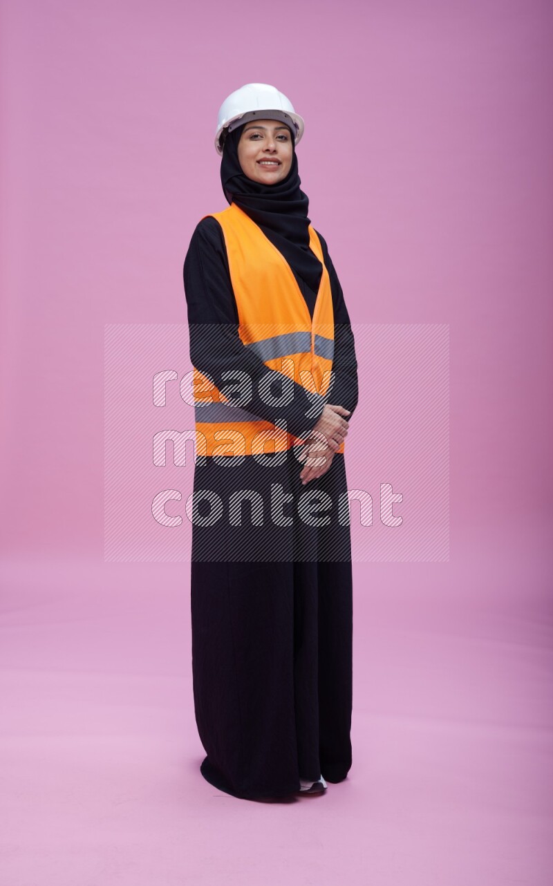 Saudi woman wearing Abaya with engineer vest and helmet standing interacting with the camera on pink background