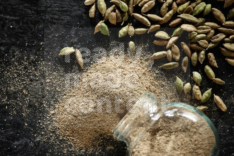 A flipped glass spice jar full of cardamom powder and powder spilled out of it with cardamom seeds spreaded on textured black flooring