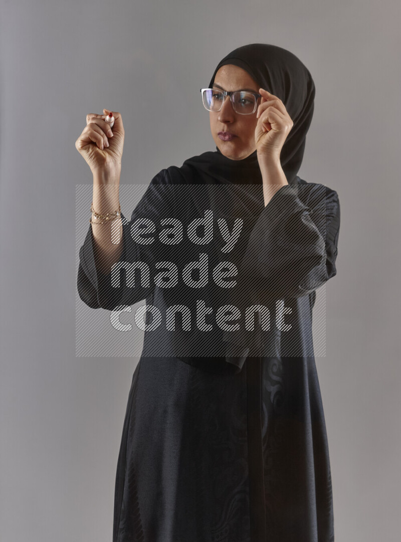 Woman in black abaya, hijab and glasses holding a marker pen to write on a transparent board with different reactions and expressions on grey background