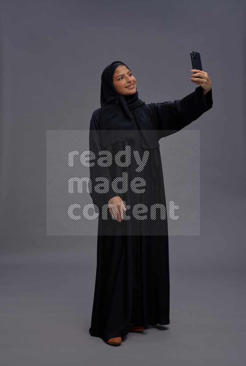 Saudi woman wearing Abaya standing taking selfie on gray background