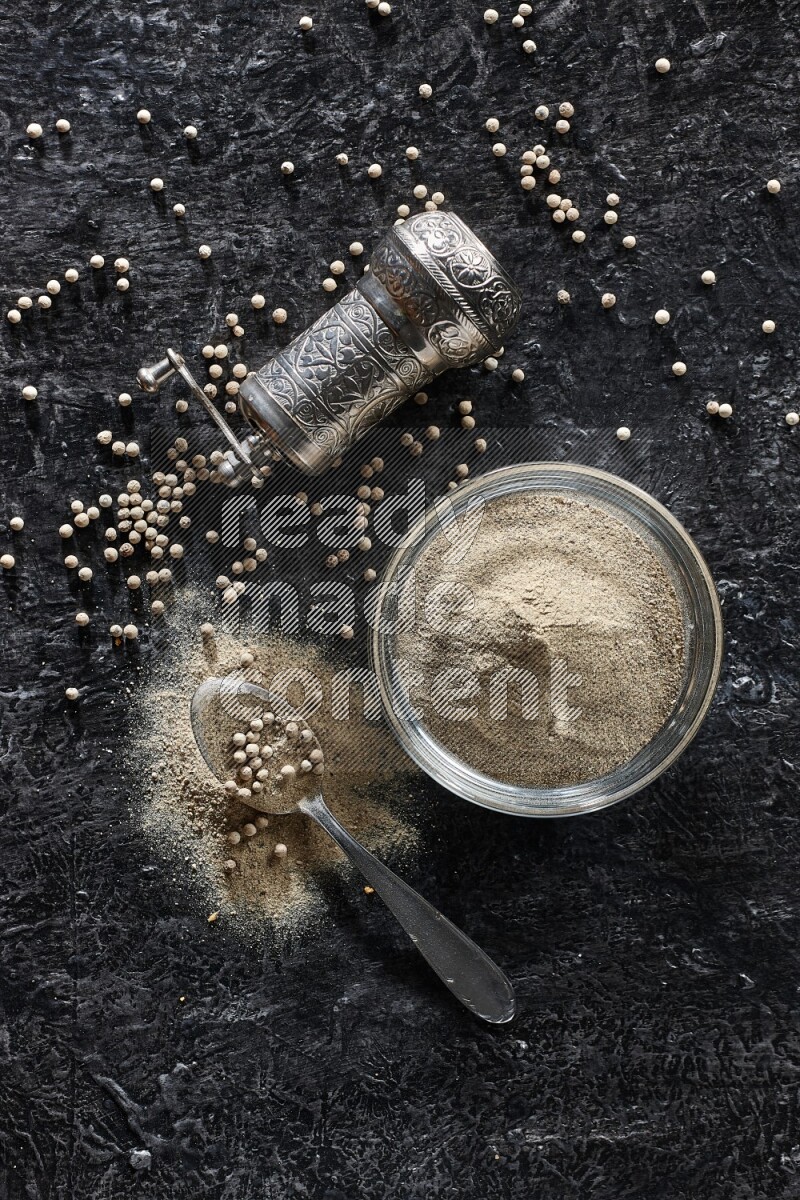 A glass bowl full of white pepper powder with pepper beads, a metal grinder and a metal spoon on textured black flooring