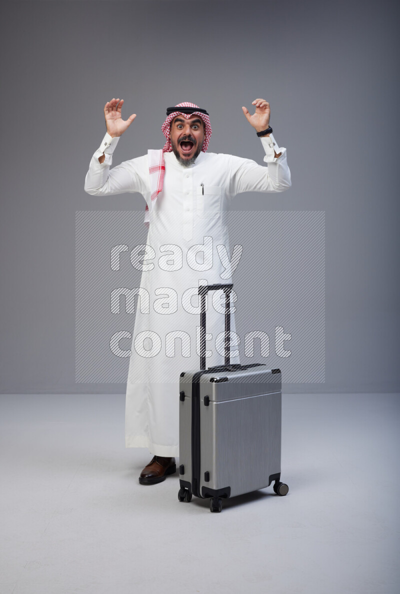 Saudi man wearing Thob and red Shomag standing holding Travel bag on Gray background