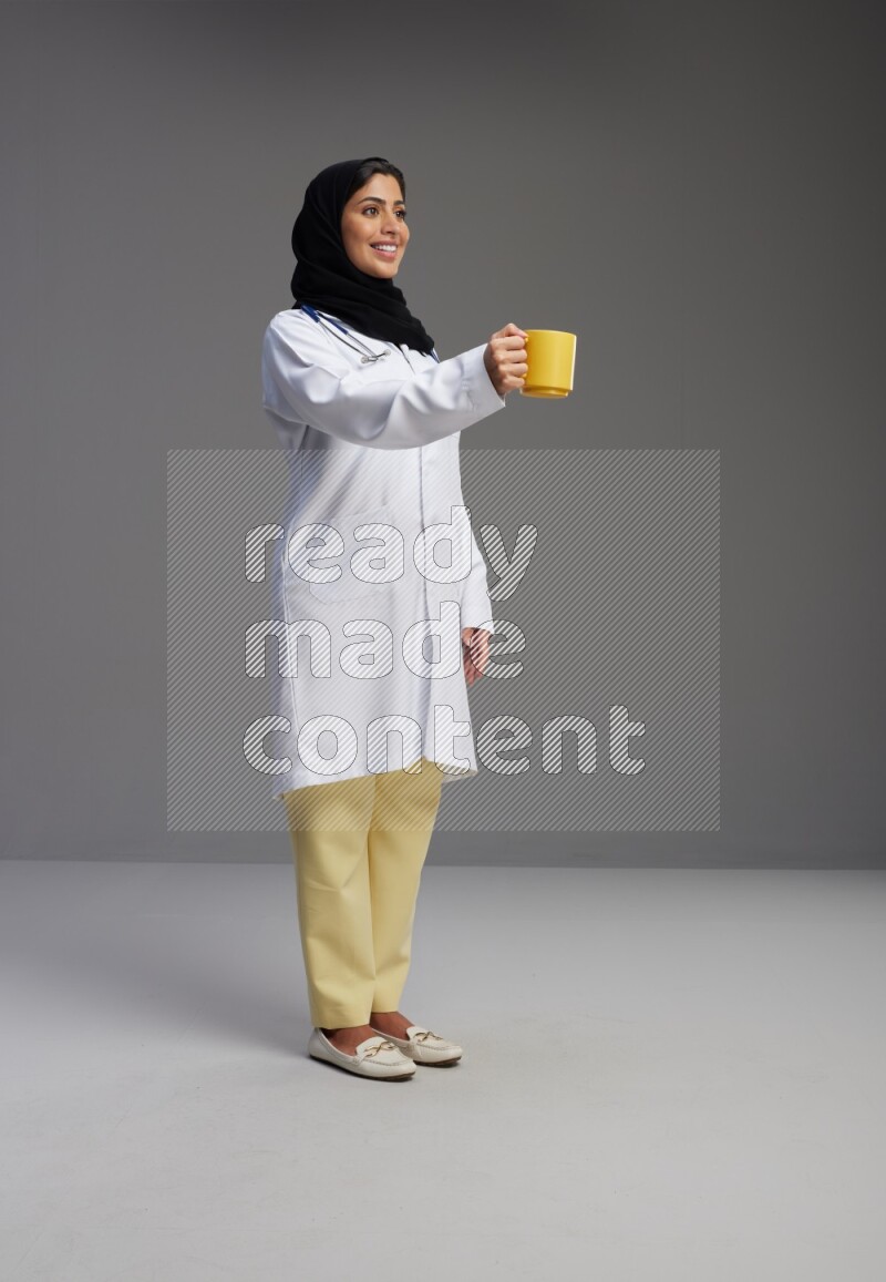 Saudi woman wearing lab coat with stethoscope standing holding mug on Gray background