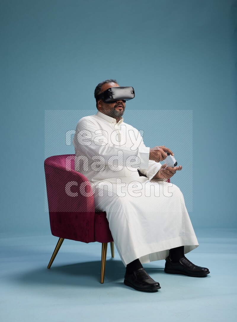 Saudi Man without shimag sitting on chair  gaming with VR and joystick on blue background